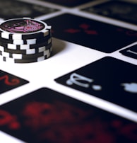 A stack of poker chips with a distinctive black and white pattern is placed on a table covered with several playing cards. The cards are dark with red and white symbols, creating a bold contrast against the lighter surface.